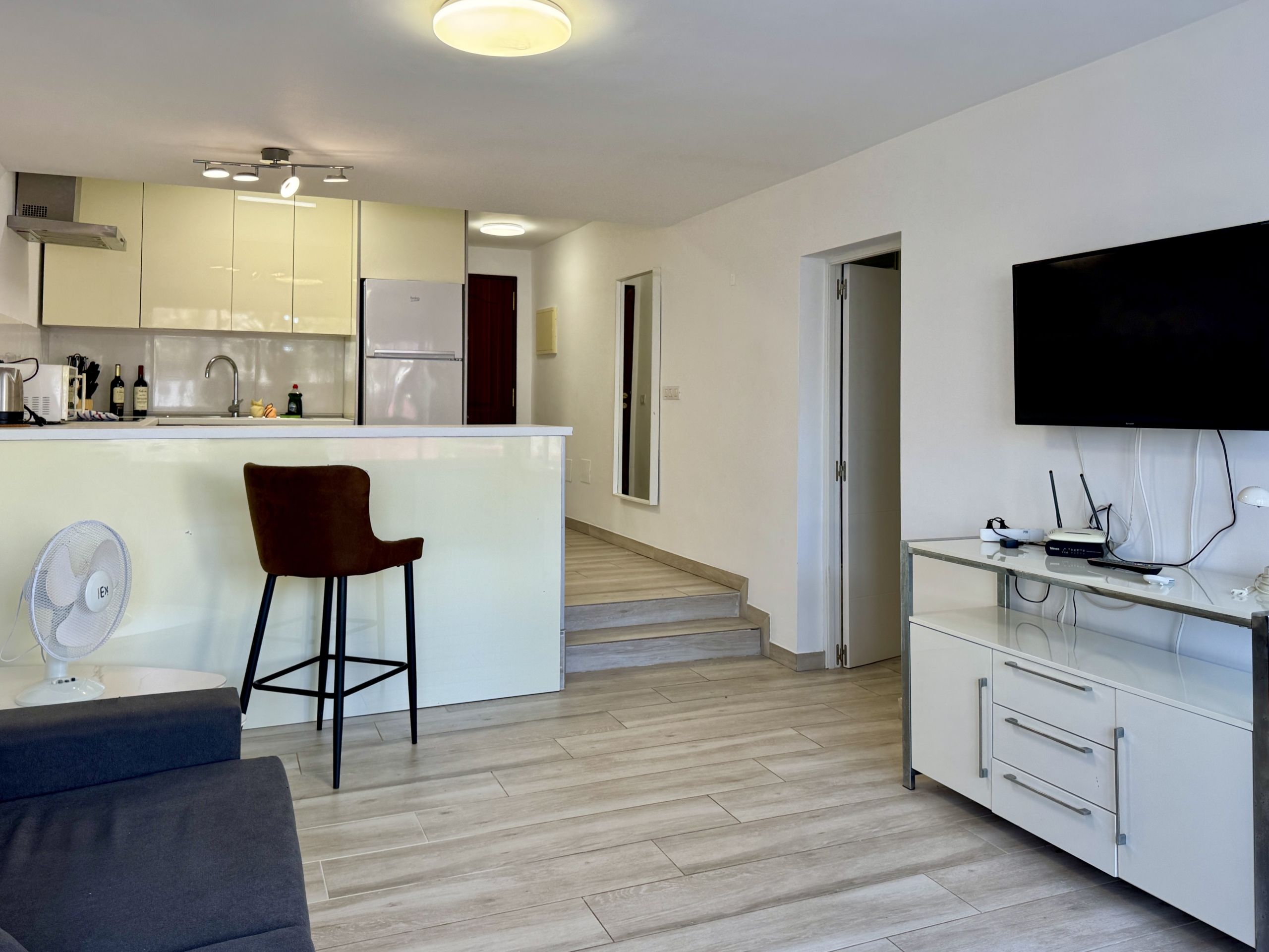 Open-plan living area with a white kitchen, bar-height stool, and a flat-screen TV on the wall to the right.