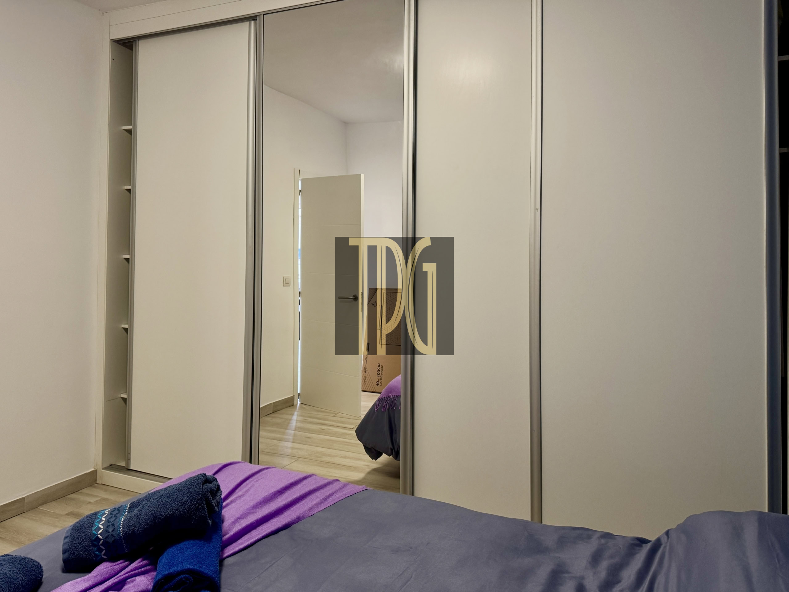 Bedroom with a large mirrored sliding wardrobe; open left section reveals shelves, bed with purple and blue towels in foreground.