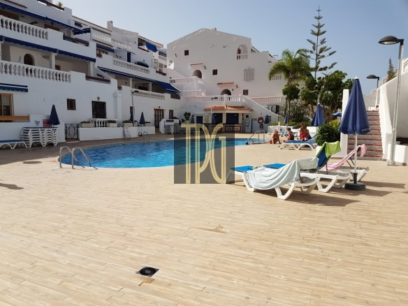 Outdoor hotel pool area with white Mediterranean buildings, blue awnings, and sun loungers around a blue pool, under a clear sky.