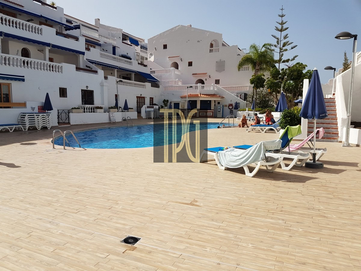 Outdoor hotel pool area with white Mediterranean buildings, blue awnings, and sun loungers around a blue pool, under a clear sky.