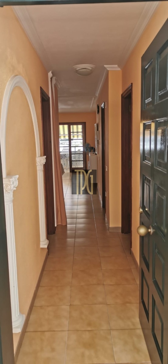 Bright hallway with beige walls, white decorative arch on the left, brown doors along the corridor, and a sunlit room at the far end.