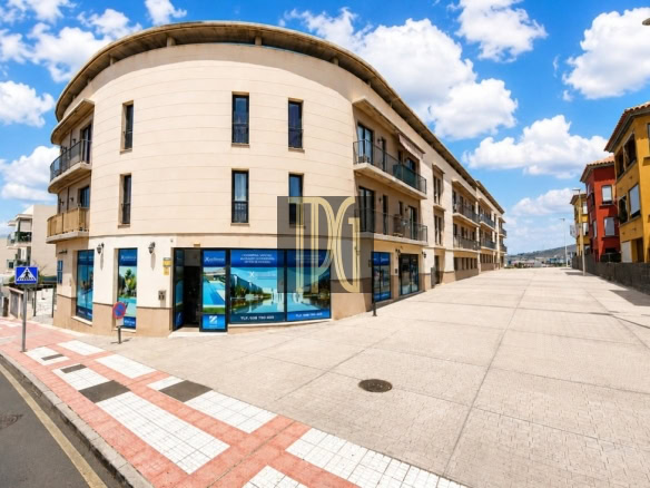 Wide street scene featuring a curved beige building with shopfronts on the ground floor and balconies above, under a blue sky with clouds.",