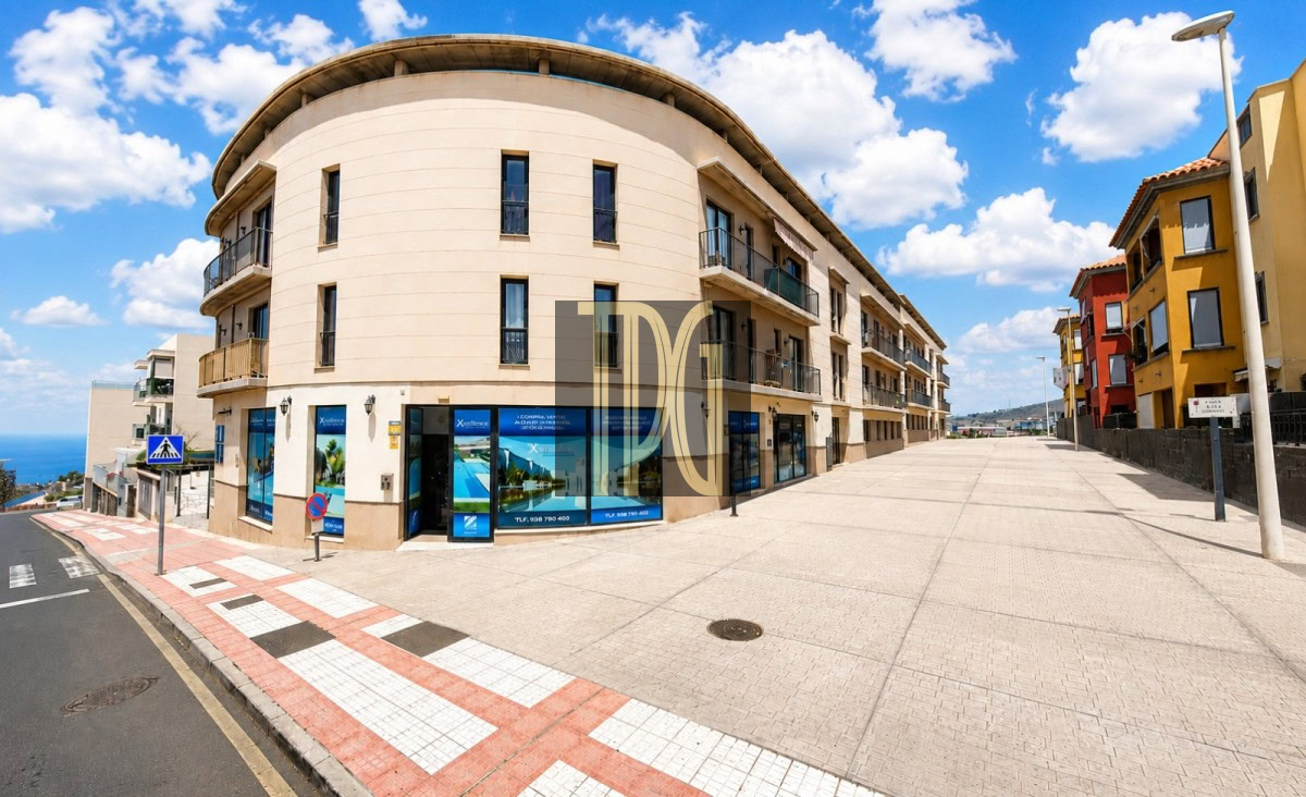 Unknown-1 Wide street scene featuring a curved beige building with shopfronts on the ground floor and balconies above, under a blue sky with clouds.",
