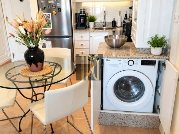 Small kitchen scene with a glass round dining table, white chairs, and a vase of dried flowers; fridge and counter area in the background.