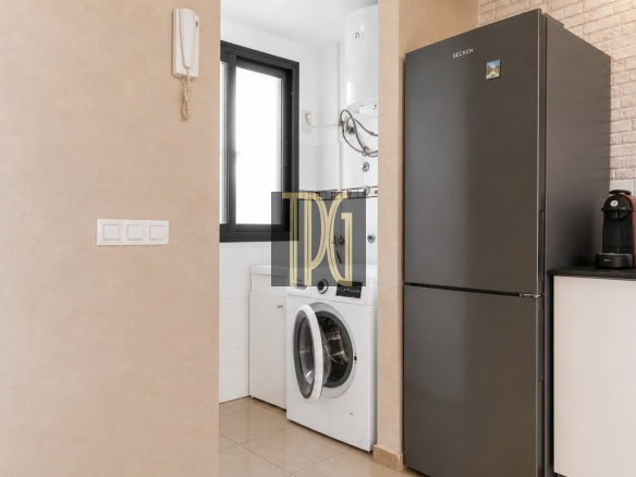 Modern laundry alcove with front-loading washing machine under a window, adjacent to a tall dark-gray refrigerator and a coffee machine on the counter.