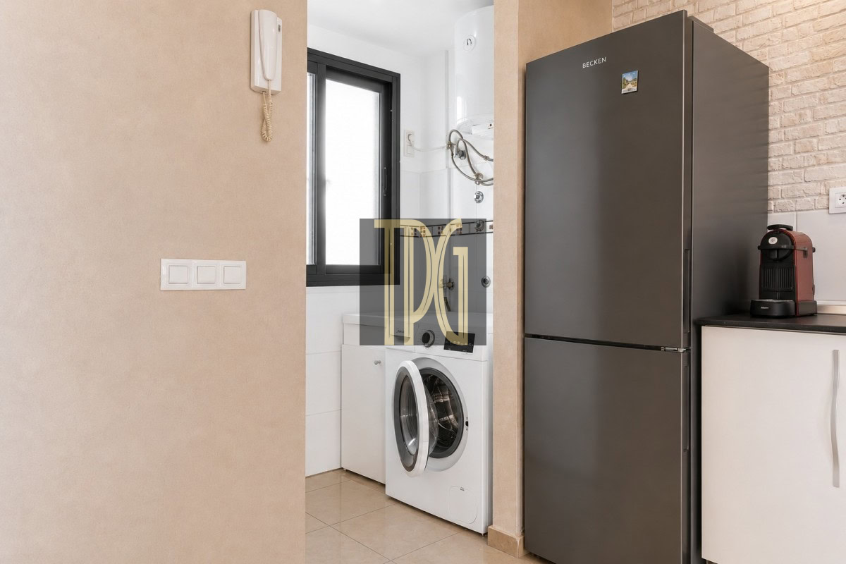 Unknown-10 Modern laundry alcove with front-loading washing machine under a window, adjacent to a tall dark-gray refrigerator and a coffee machine on the counter.