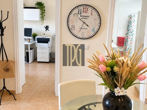 Round vintage clock on beige wall overlooking a glass dining table with a floral arrangement, and a doorway to a home office with a printer in the background