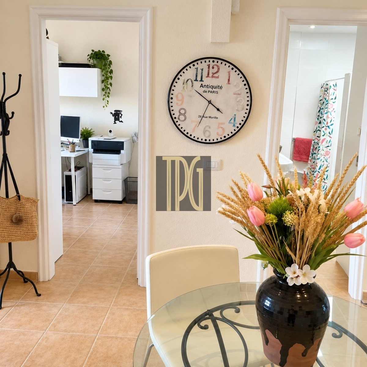 Unknown Round vintage clock on beige wall overlooking a glass dining table with a floral arrangement, and a doorway to a home office with a printer in the background