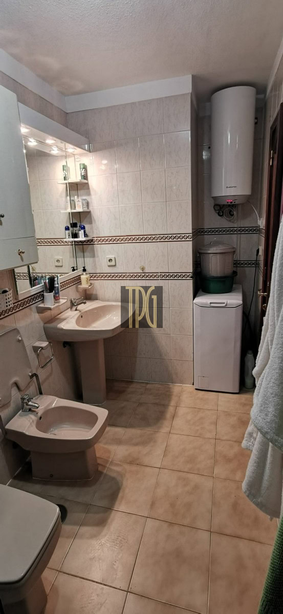 Small beige-tiled bathroom with toilet, bidet, pedestal sink, mirror cabinet, and a washing machine under a wall-mounted water heater.