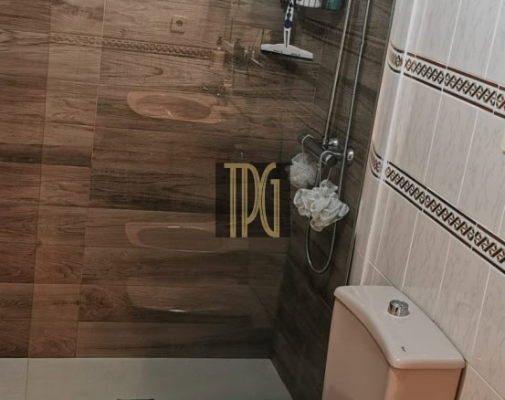 Beige toilet and bidet beside a glass-enclosed shower with chrome fixtures and wall shelves holding shampoo bottles in a tiled bathroom.