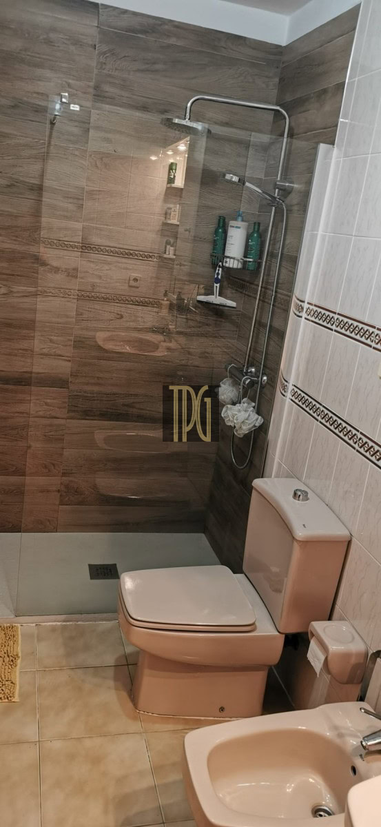 Beige toilet and bidet beside a glass-enclosed shower with chrome fixtures and wall shelves holding shampoo bottles in a tiled bathroom.