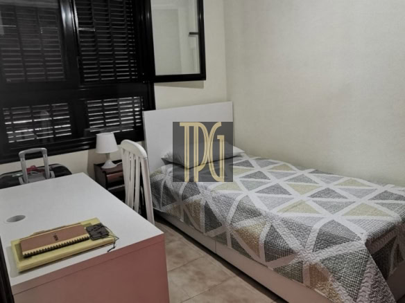 Small bedroom with a single bed covered in a geometric quilt, a white desk and chair near a window with dark blinds.