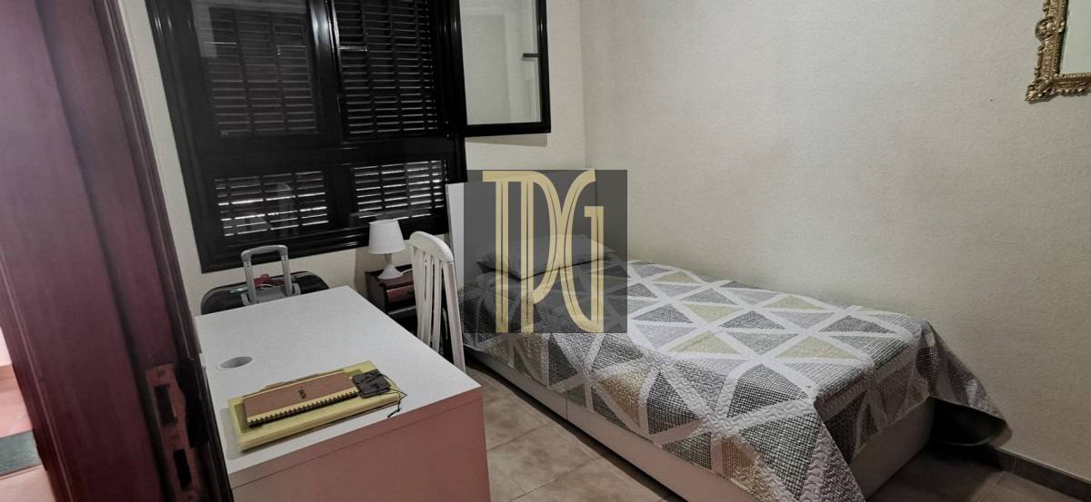 Small bedroom with a single bed covered in a geometric quilt, a white desk and chair near a window with dark blinds.