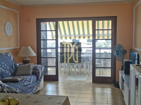 Living room with orange walls, blue patterned sofa, and a glass dining table; balcony doors open to a striped awning patio outside edge of view.