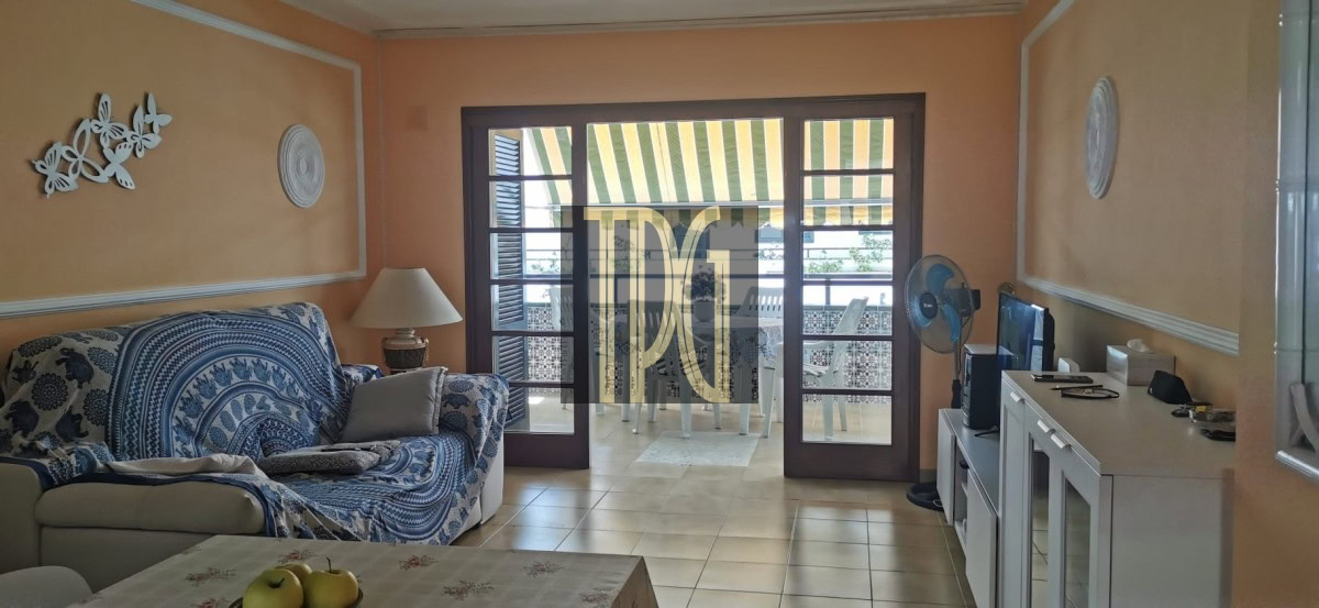 Living room with orange walls, blue patterned sofa, and a glass dining table; balcony doors open to a striped awning patio outside edge of view.