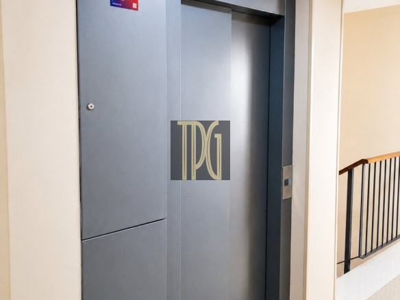 Grey elevator doors closed in a beige-halled corridor, with a railing on the right side and tiled floor.
