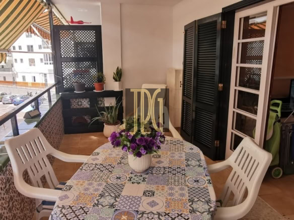 Balcony dining area with a mosaic table, white plastic chairs, striped awning, and potted plants; street view beyond railing.