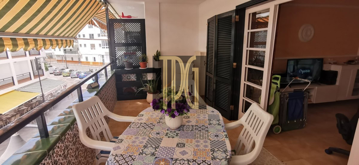 Balcony dining area with a mosaic table, white plastic chairs, striped awning, and potted plants; street view beyond railing.
