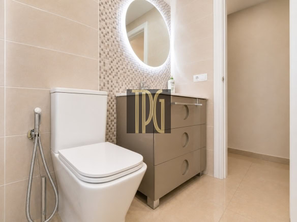 Modern beige bathroom with a wall-mounted vanity, circular backlit mirror, and mosaic tile backsplash beside a white toilet and bidet sprayer