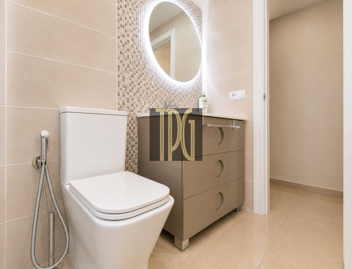 Unknown-8 Modern beige bathroom with a wall-mounted vanity, circular backlit mirror, and mosaic tile backsplash beside a white toilet and bidet sprayer