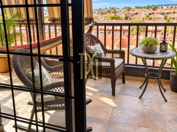 Balcony seating with a wicker chair and round table, potted plant and lantern, overlooking a sunny neighborhood of red-tile roofs.