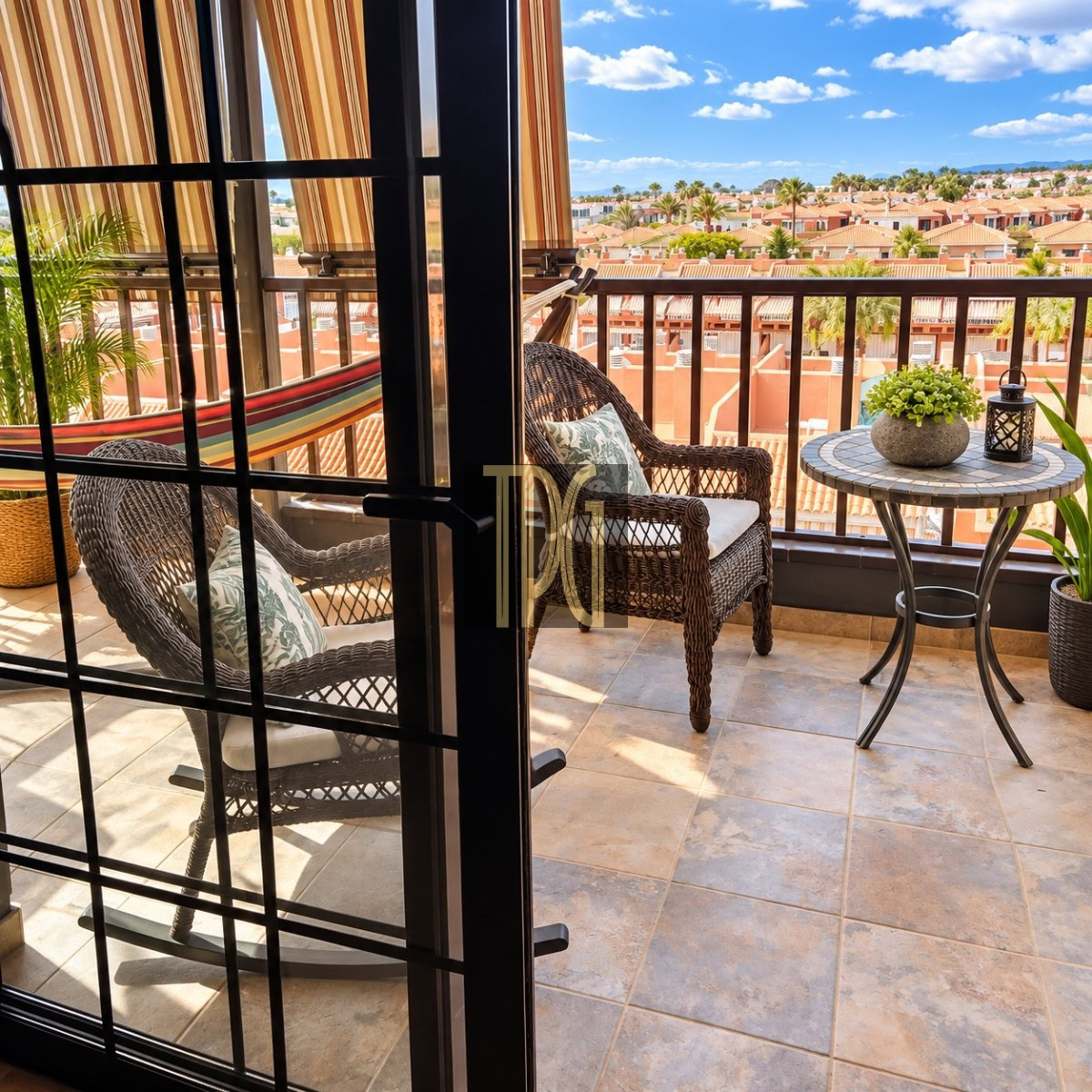 Unknown-9 Balcony seating with a wicker chair and round table, potted plant and lantern, overlooking a sunny neighborhood of red-tile roofs.