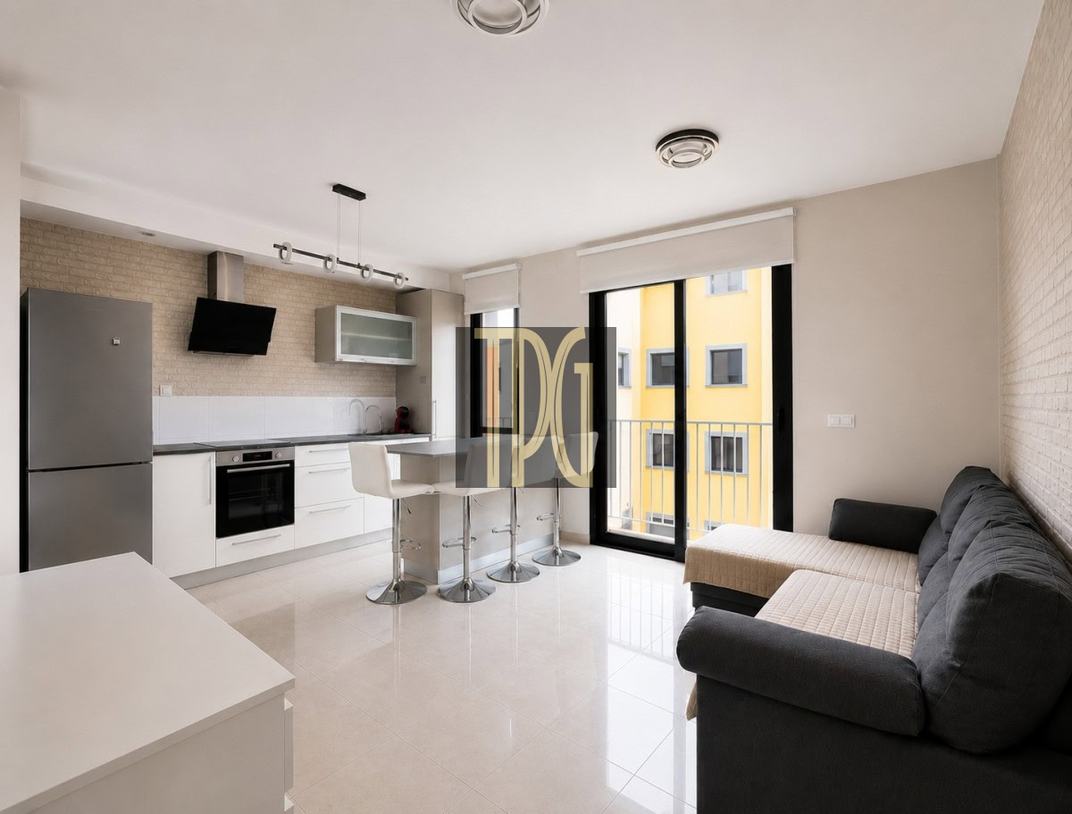 Unknown-9 Open-plan kitchen with white cabinets, an island with bar stools, and a gray sofa facing a balcony door.